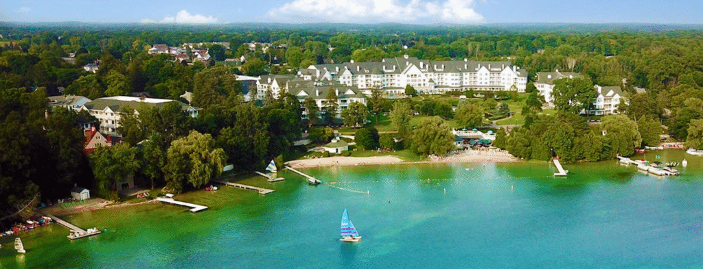 A scenic aerial view of Elkhart Lake, Wisconsin, showing turquoise lake water, sailboats, sandy beaches, lush green trees, and the white resort buildings along the shoreline under a blue sky.
